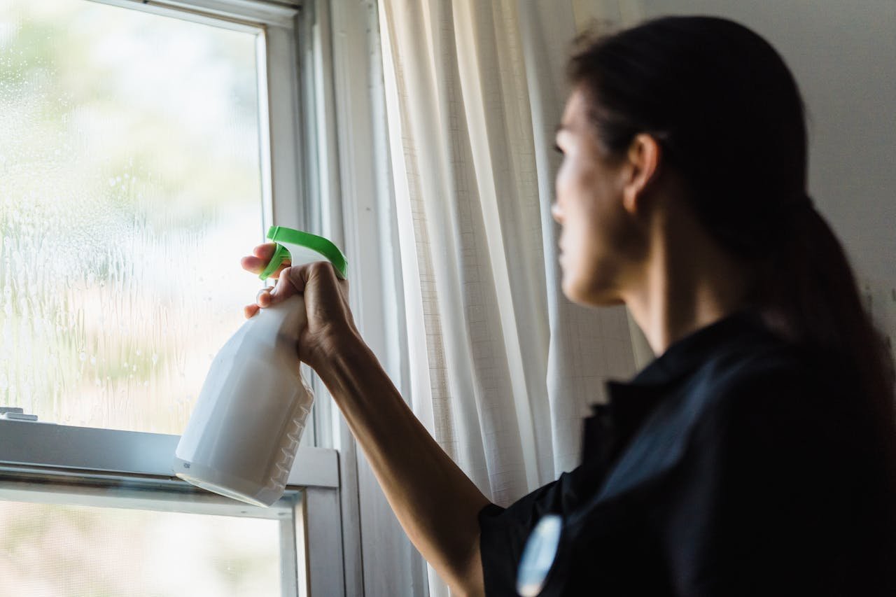 A woman using a spray bottle to clean a glass window inside a room, captured in a close-up shot.